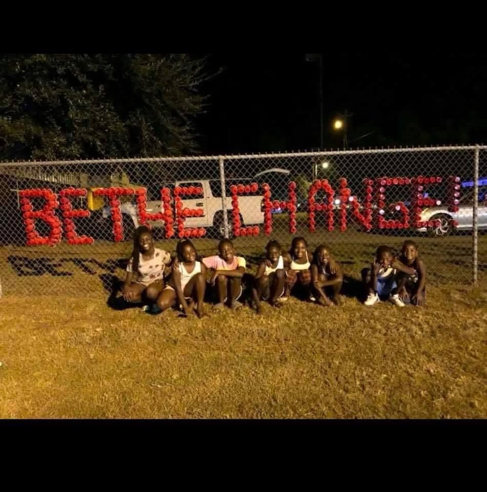 Children at night in front of a fence with BE THE CHANGE spelled in red cups