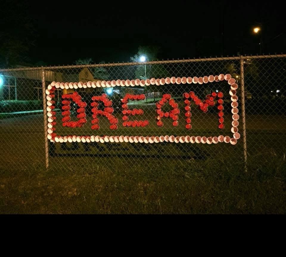DREAM spelled in red cups inside a white cup border on a fence at night