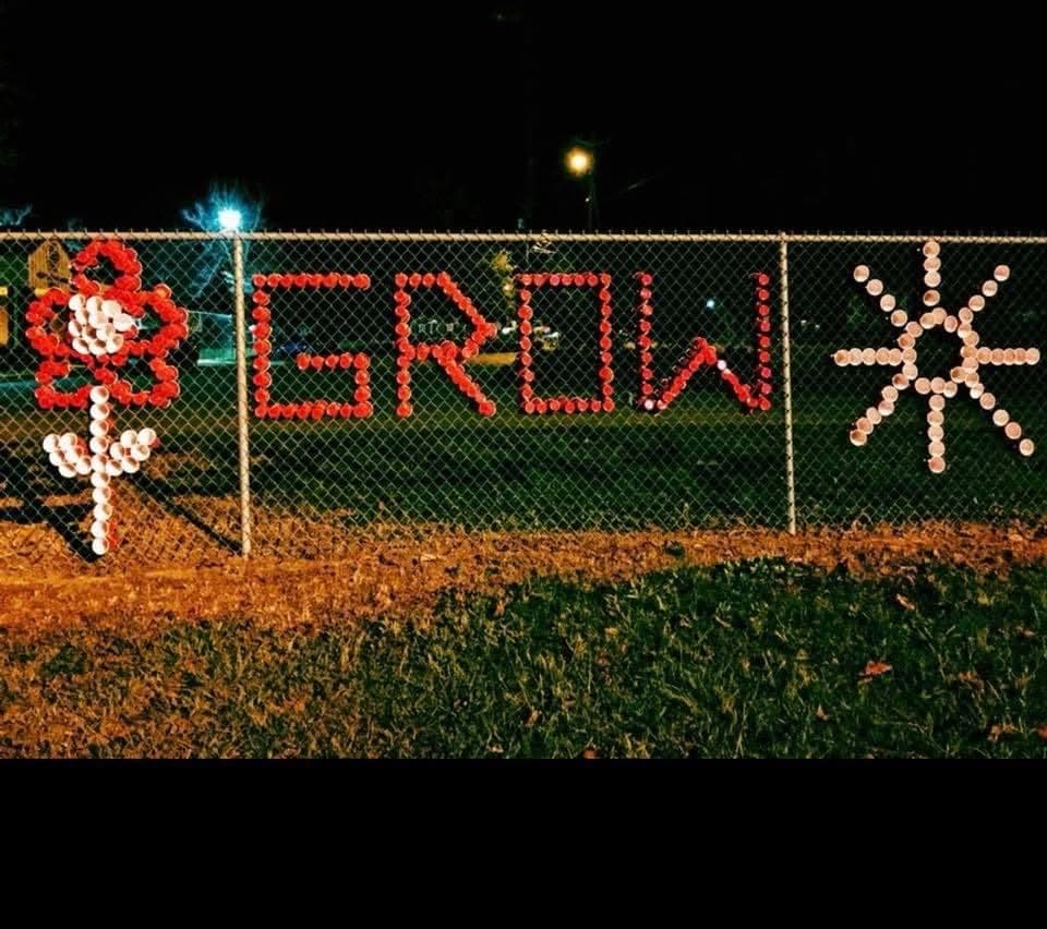 GROW with flower and sun symbols spelled in red and white cups on a fence at night